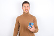 © Jihan - young caucasian man wearing knitted turtleneck over white background holds mobile phone in hands and rejoices positive news, uses modern cellular