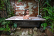 A young woman looking over her shoulder smiles happily while sitting in an outdoor bubble bath in a lush tropical garden.  