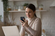 © fizkes - Attractive millennial woman use cellphone seated at table with laptop in cozy kitchen. Young generation and modern wireless tech usage or fun and business remotely from home. E-services user concept