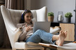 Young woman in headphones relax on cozy armchair put feet on footstool, look at laptop screen, talk to friend, share news to family living abroad through video conference app. Virtual meeting concept