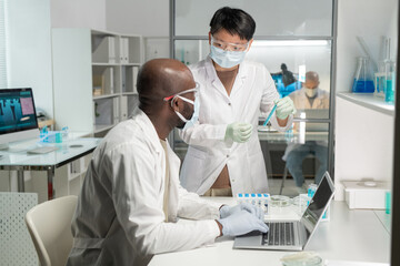 Young Asian female scientist in protective mask and lab coat showing her African American male colleague flask with blue liquid