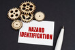 On the black surface are gears, a pen and a business card with the inscription - Hazard Identification