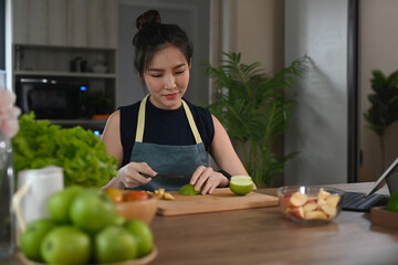 Wall Mural - Healthy young woman sitting at kitchen counter and cutting fresh organic apple on wooden board.