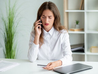 Bank payment. Worried woman. Mobile consultation. Considered office lady holding credit card talking smartphone sitting work desk in light room interior.