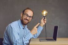 Young happy clever caucasian business man freelancer holding illuminated lightbulb in hand and pointing with forefinger sitting front of laptop at desk. Idea, brainstorming process, creative thinking