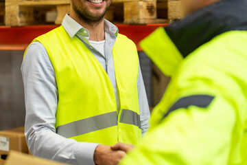 Wall Mural - wholesale, logistic, people, agreement and export concept - close up of manual worker and businessmen in reflective safety vests shaking hands and making deal at warehouse