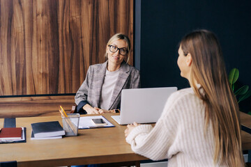 Two young modern business women smiling talking while sitting at the desk in the office.