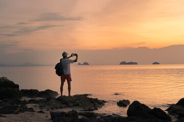 Wall Mural - A man on the beach shoots a colorful sunset on a smartphone.Spectacular sunset sky over sea and islands.