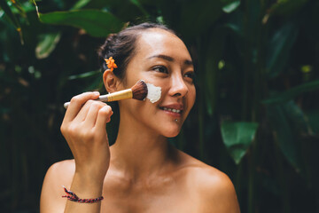 Happy female apply scrub cosmetic on face skin using makeup brush while doing beauty procedures in nature environment enjoying healthy lifestyle, pretty woman with organic cream smiling and relaxing