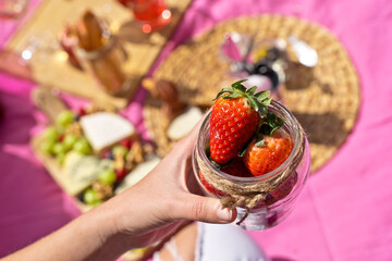 Wall Mural - Horizontal top view of unrecognizable woman holding a bowl of strawberries outdoors. Above shot of woman holding strawberries on pink picnic background. Health and food concept.