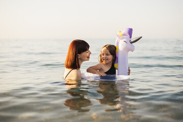 Wall Mural - Side view of happy caucasian mother teaching swimming little lovely smiling daughter with white inflatable lifebuoy unicorn in sea water. Smiling family having fun, summertime outdoors concept.