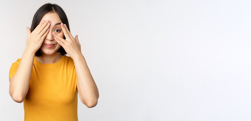 Cute korean female model, cover eyes with hands, peeking through fingers curious, watching smth, standing in yellow tshirt over white background