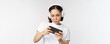 Happy asian woman in headphones, looking at smartphone, watching video on mobile phone and smiling, standing over white background