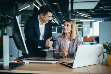 Wall Mural - Two employees in a modern office, an Asian man and a woman working at a table, colleagues discussing and consulting, thinking about a joint project