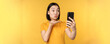 Image of happy, beautiful asian girl video chatting, talking on smartphone application, standing against yellow background