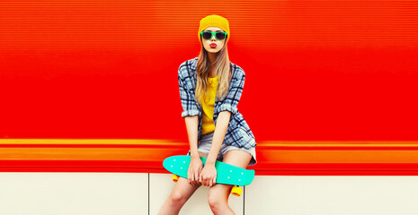 Poster - Portrait of stylish blonde young woman model with skateboard wearing colorful yellow hat on red background