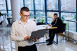 © Дмитрий Ткачук - A young businessman man in glasses and a white shirt with a laptop in his hands stands in the office and works