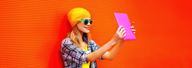 Portrait of stylish cool young woman using tablet pc with skateboard wearing colorful clothes on vivid background