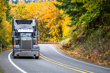 Wall Mural - Powerful old style dark gray classic big rig semi truck with refrigerator unit on the front wall of the reefer semi trailer running on the winding mountain road through the autumn forest