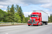 Stylish Bright Red Big Rig Semi Truck With Chrome Parts Transporting Cargo In Dry Van Semi Trailer Running On The Straight Wide Highway Road With Green Trees On The Side