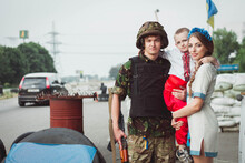 Young Ukrainian Woman And Child Boy Stand Near Ukrainian Soldier On Roadblock Against Background Of Sandbags.