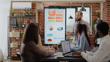 Wall Mural - Worker talking about business growth with charts on monitor, doing financial presentation for workmates in office. Person meeting with people to plan project and startegy for development.