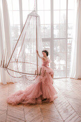 young woman with short black hair in pink princess dress with train is sitting on the crystal chandelier near huge panoramic window background like dior style. fashion concept, free space