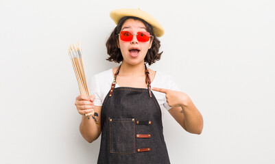 Wall Mural - pretty hispanic girl looking shocked and surprised with mouth wide open, pointing to self. art student concept