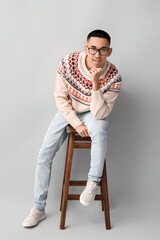 Wall Mural - Handsome young Asian man in knitted sweater sitting on chair against grey background
