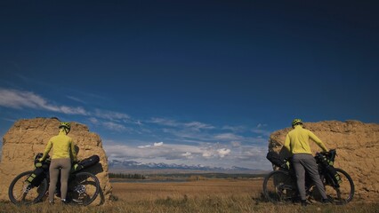 The man and woman travel on mixed terrain cycle bike touring with bikepacking. The two people journey with bicycle bags. Sport sportswear in green black colors. Mountain snow capped, stone arch.