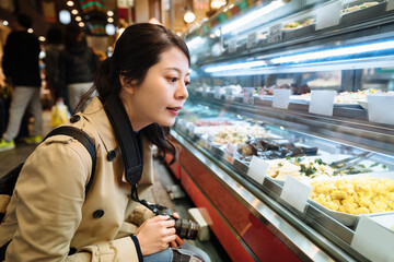 woman backpacker carrying camera is looking at variety of gourmet in deli section in Japan. chinese girl holding digital camera is selecting from collection of food displayed in a showcase.