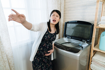 Wall Mural - young asian japanese pregnant woman suffering from stomach ache at home while doing house chores. sickness maternity lady holding big belly with painful feeling in laundry room at apartment indoors.