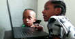 © Marco - African teen girl browsing internet on laptop, little brother watching next to sibling