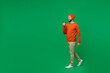 Full body side view fun young smiling happy man 20s wear orange sweatshirt hat hold takeaway delivery craft paper brown cup coffee to go walk stroll isolated on plain green background studio portrait