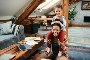 Happy black mother having fun while carrying her daughter on shoulders at home.