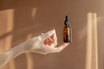 Wall Mural - Woman's hand holds an amber glass bottle with dropper cap on brown background. Unmarked bottle with serum for face, cosmetic product. Concept of skin care and beauty