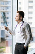 © dikushin - Vertical portrait of confident male doctor in white uniform examining chest x-ray lungs scan standing near window in hospital office, thinking about diagnosis. Concept of medicine and health care.
