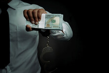 Young Businessman Standing Holding Money And Handcuffs Attached To His Arm Concept Corrupt Police, Bad Cop, Bribe Police.