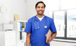 healthcare, profession and medicine concept - happy smiling doctor or male nurse in blue uniform with clipboard and stethoscope over medical office at hospital background