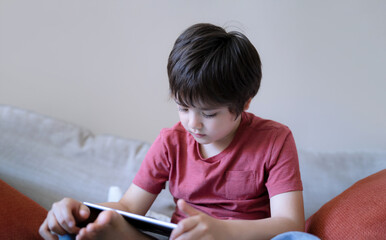 Wall Mural - School kid sitting on sofa watching cartoon on tablet, Cute young boy playing game online on digital pad, Cinematic indoor portrait Child having fun and relaxing on his own in study room