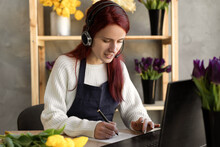 Young Female Florist In Blue Apron Business Owner Using Headset And Computer While Talking To Customers Online Taking Order Online.