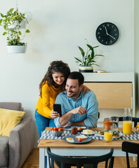 Wall Mural - Happy Couple Using Mobile Phone Together While Eating Breakfast at Home