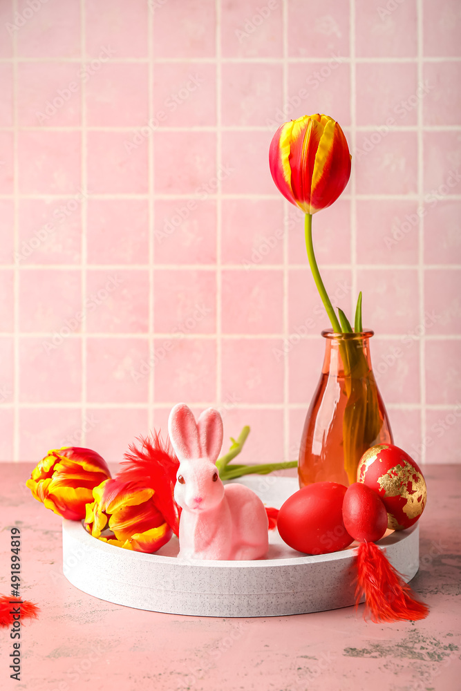 Stand with cute Easter bunny, painted eggs and flowers on pink background