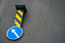 A Road Sign With A White Arrow On A Blue Background Lies On The Roadway. A Strong Wind Knocked Over A Road Sign.