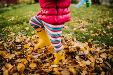 Toddler Girl Stomps Through Pile Of Leaves In Yellow Boots