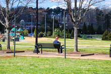 A Woman In A Red Hat Sitting On A Black Park Bench Surrounded By Bare Winter Trees, Tall Black Light Posts Green And Yellow Grass With Blue Sky And Clouds At Swift Cantrell Park In Kennesaw Georgia