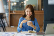 © amnaj - Charming Asian woman with a smile sitting holding smartphone at the office.