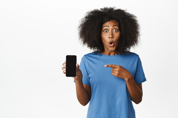 Wall Mural - Portrait of amazed and surprised Black woman pointing finger at mobile phone screen, showing smartphone application with enthusiastic face, white background