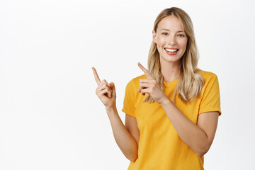 Wall Mural - Portrait of young woman demonstrating, showing announcement or logo, pointing fingers left, standing in yellow t-shirt over white background