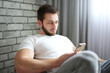 © Denis Barysau - A young man of athletic build in a white T-shirt sits on a cushion chair and looks intently intosmartphone
